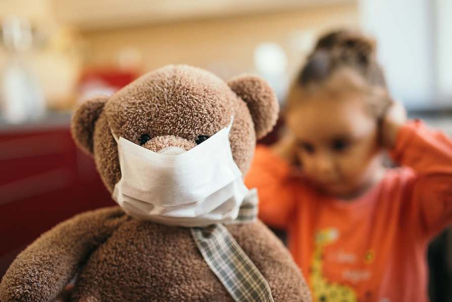 Teddy bear wearing a face mask in front of a girl with hands on her head.